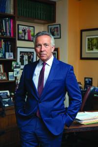 A man in a blue suit leans against a desk with his hands in his pockets